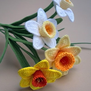May include: Crocheted daffodils in white, yellow, and orange with green stems and leaves. The handmade flowers have detailed petals and trumpet-shaped centers, creating a realistic floral design.