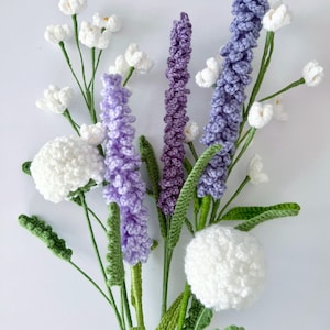Puede incluir: Un ramo de flores de ganchillo hecho a mano. El arreglo presenta flores de color lavanda, moradas y blancas con tallos y hojas verdes. Las flores están dispuestas sobre un fondo blanco.