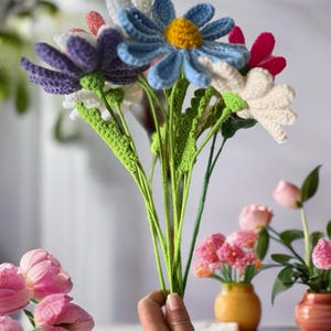 Op de afbeelding: Een boeket gehaakte bloemen in verschillende kleuren, waaronder paars, blauw, wit en roze. De bloemen zijn in een hand gerangschikt, met de stelen die naar beneden reiken. De bloemen zijn gemaakt van garen en hebben een realistisch uiterlijk.