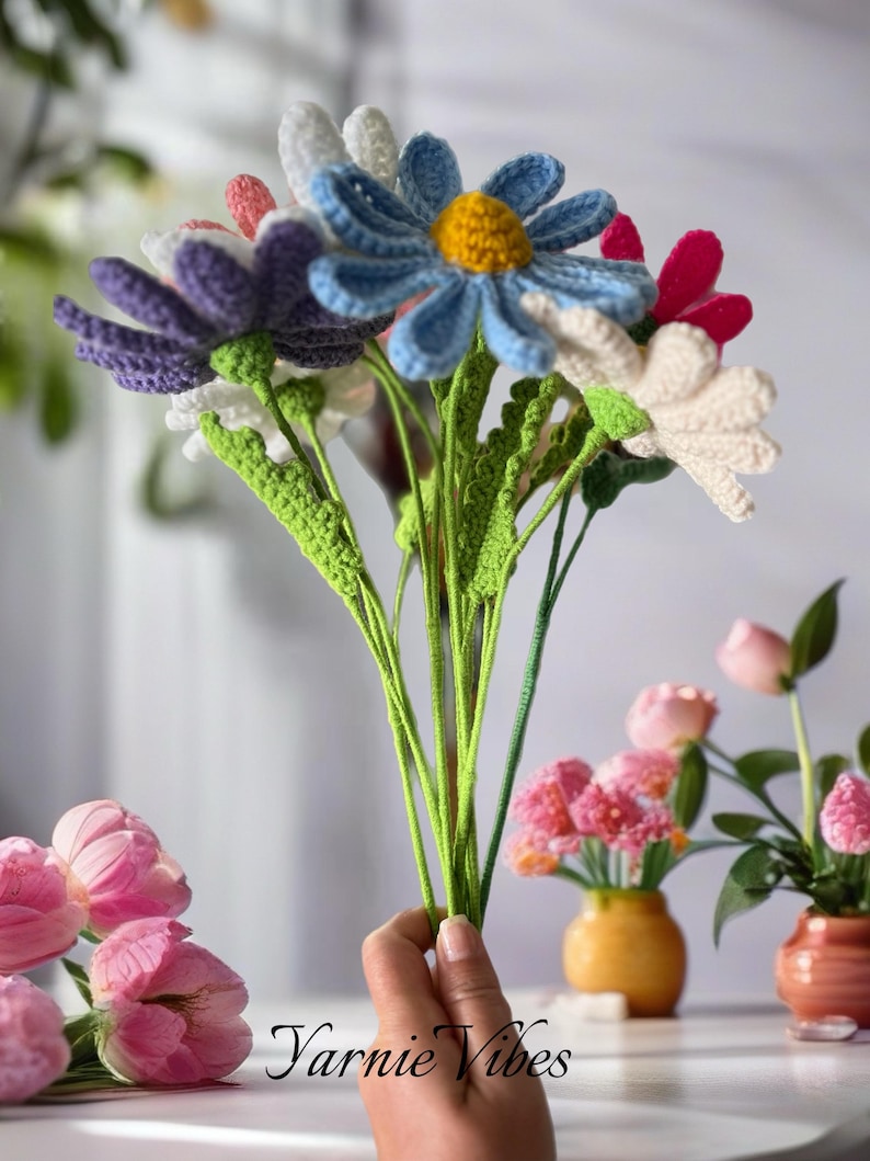 Op de afbeelding: Een boeket gehaakte bloemen in verschillende kleuren, waaronder paars, blauw, wit en roze. De bloemen zijn in een hand gerangschikt, met de stelen die naar beneden reiken. De bloemen zijn gemaakt van garen en hebben een realistisch uiterlijk.