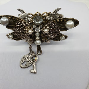 May include: A bronze dragonfly brooch with pearl accents and a silver key charm with a heart-shaped keyhole. The keyhole has the word "Dior" inscribed on it.