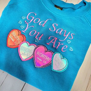 May include: A turquoise sweatshirt with the embroidered phrase "God Says You Are" in pink. Below the text are heart-shaped patches with words like "Beautiful", "Strong", "Worthy", and "Loved".