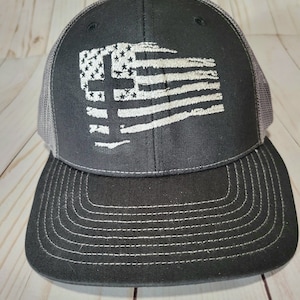 May include: A black and gray trucker hat with a white distressed American flag design featuring a cross in the center.