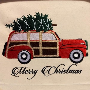 May include: A red vintage station wagon with a wooden panel design is carrying a green Christmas tree on top. The words "Merry Christmas" are embroidered below the car.