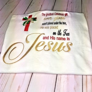 May include: White t-shirt with gold embroidery that reads "The greatest Christmas gift ever given wasn't placed under the tree, He was placed on the tree and His name is Jesus". A red embroidered cross with holly and a bow is in the top left corner.