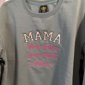 May include: A teal sweatshirt with the word "MAMA" embroidered in pink and green floral letters. Below the word "MAMA" is the text "Strong Fearless Loving Patient Selfless" embroidered in pink.