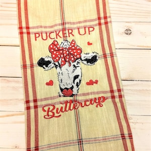 May include: A beige kitchen towel with red and black plaid pattern. The towel features an embroidered cow with a red bow and the text "Pucker Up Buttercup".