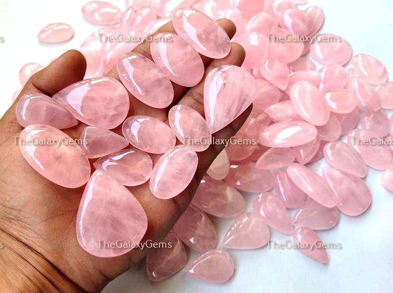 May include: A large collection of pink rose quartz cabochons in various sizes and shapes. The cabochons are smooth and polished, with a translucent appearance. The Galaxy Gems logo is visible in the image.