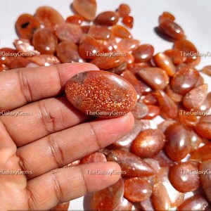 May include: A collection of sunstone gemstones in various shapes and sizes. The stones are a mix of orange, red, and gold colors with a shimmery, iridescent finish.