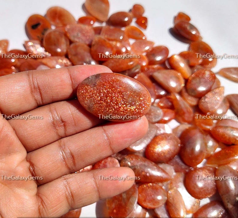 May include: A collection of sunstone gemstones in various shapes and sizes. The stones are a mix of orange, red, and gold colors with a shimmery, iridescent finish.