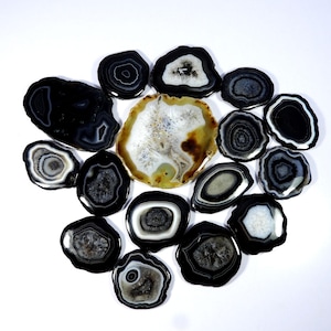 May include: A selection of polished agate slices in various shapes and sizes, arranged on a white surface. The slices display concentric bands of black, white, and grey, with some featuring brown and cream-coloured centres. The overall theme is natural stone decor.