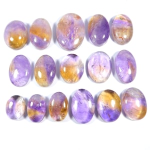 May include: A collection of oval-shaped gemstones with a mix of purple and golden-yellow hues. The stones are polished and smooth, with variations in color and pattern. The gemstones are arranged on a white surface.