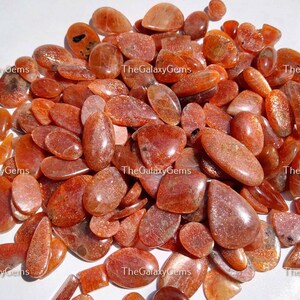 May include: A large pile of orange sunstone cabochons, some with a pear shape, others oval, and some with a more rounded shape. The stones are translucent and have a shimmery, glittery appearance.