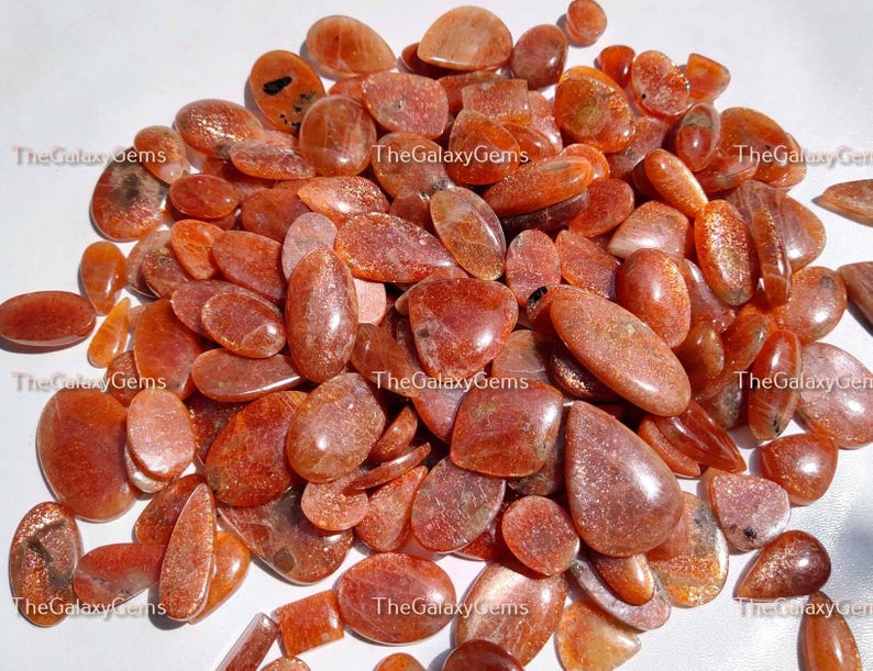 May include: A large pile of orange sunstone cabochons, some with a pear shape, others oval, and some with a more rounded shape. The stones are translucent and have a shimmery, glittery appearance.