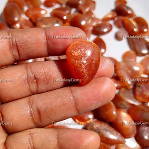 May include: A close-up of a hand holding a heart-shaped sunstone gemstone. The gemstone is a deep orange color with a shimmery, iridescent finish. The background is a pile of similar sunstone gemstones.