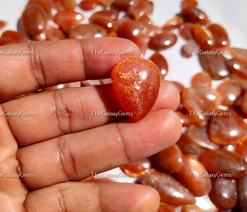 May include: A close-up of a hand holding a heart-shaped sunstone gemstone. The gemstone is a deep orange color with a shimmery, iridescent finish. The background is a pile of similar sunstone gemstones.