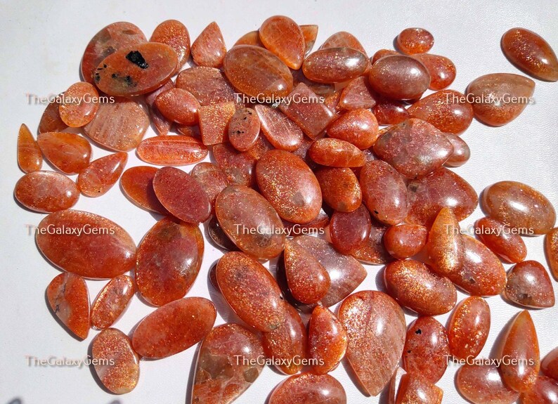 May include: A collection of approximately 40 sunstone cabochons in various shapes and sizes. The stones are a mix of orange, red, and brown with a shimmery, iridescent quality.