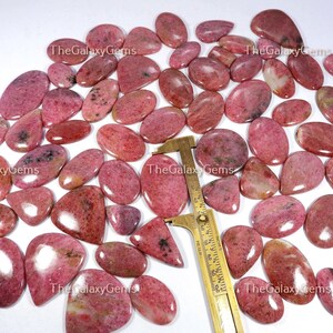 May include: A collection of 30 pink and white gemstone cabochons, each with a unique pattern and shape. The cabochons are arranged on a white surface with a ruler for scale. The cabochons are approximately 2.5 cm in length.