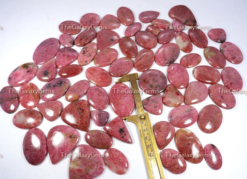 May include: A collection of 30 pink and white gemstone cabochons, each with a unique pattern and shape. The cabochons are arranged on a white surface with a ruler for scale. The cabochons are approximately 2.5 cm in length.