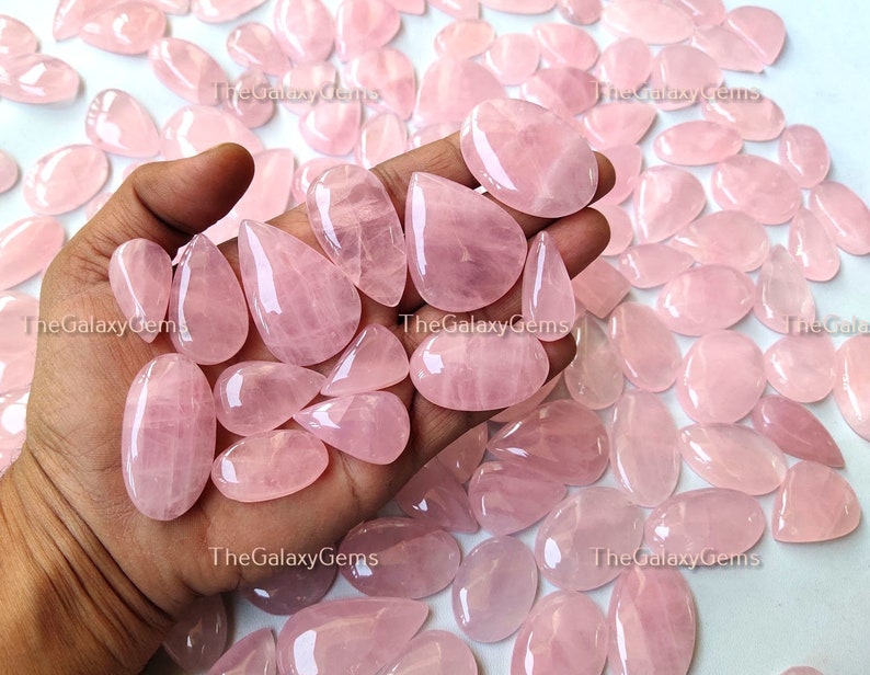May include: A collection of pink rose quartz cabochons, some with a teardrop shape, others with a more oval shape. The stones are smooth and polished, with a translucent appearance.