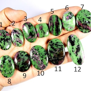 Peut inclure: Une collection de douze pierres précieuses de zoïsite polies, chacune avec un motif unique de teintes vertes, noires et roses. Les pierres varient en forme, y compris ovale et rectangulaire, et sont présentées dans une main.