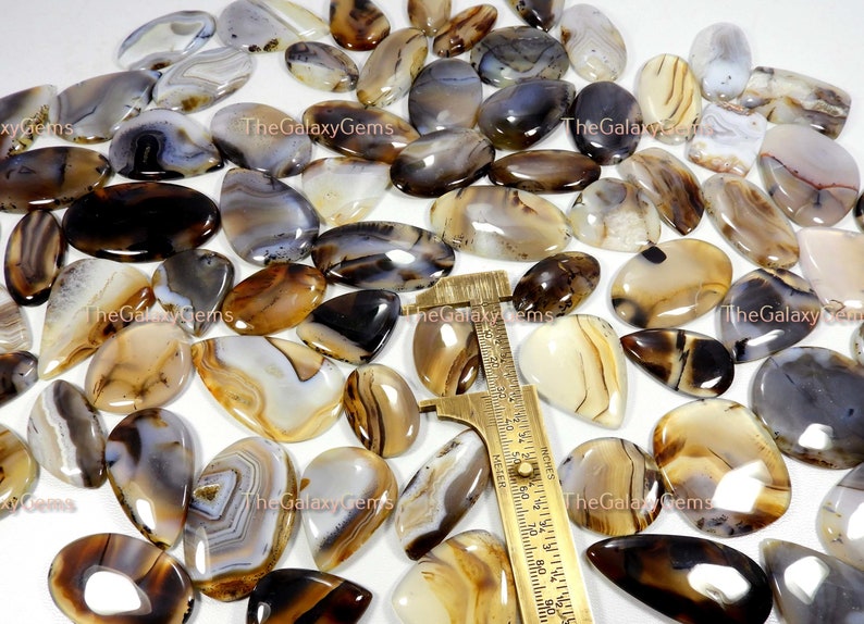 May include: A collection of agate cabochons in various shades of brown, white, and gray. The cabochons are oval-shaped and have a smooth, polished finish. The cabochons are arranged in a random pattern on a white surface.