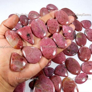 May include: A collection of pink and red gemstone cabochons, some with a pear shape and others oval, in various sizes. The stones are smooth and polished, with a translucent appearance.