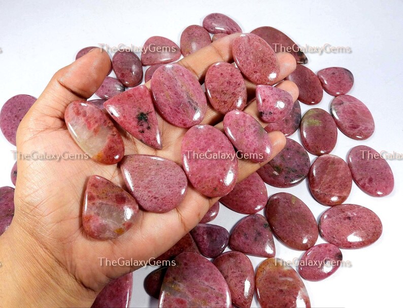 May include: A collection of pink and red gemstone cabochons, some with a pear shape and others oval, in various sizes. The stones are smooth and polished, with a translucent appearance.
