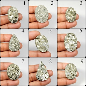 May include: Nine pyrite gemstone cabochons in various shapes, including oval and teardrop. The gemstones have a metallic, gold-like appearance with a crystalline structure. Each stone is held in a hand, showcasing the natural variations in color and texture.