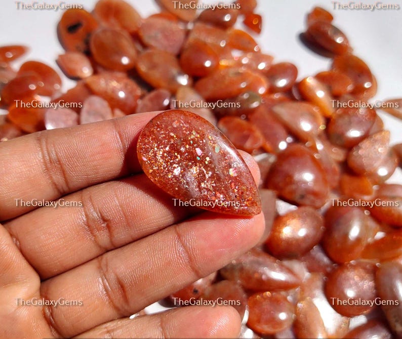 May include: A close-up of a hand holding a large, pearlescent, orange sunstone cabochon. The stone is surrounded by many smaller, orange sunstone cabochons.