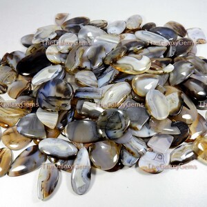 May include: A large pile of agate cabochons in various shades of brown, gray, and white. The cabochons are all oval shaped and have a smooth, polished finish.