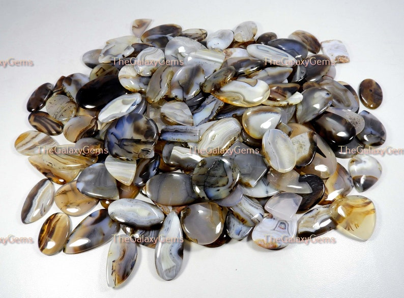May include: A large pile of agate cabochons in various shades of brown, gray, and white. The cabochons are all oval shaped and have a smooth, polished finish.