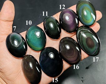 May include: A collection of oval-shaped rainbow obsidian cabochons labeled with numbers 7, 10, 11, 12, 13, 14, 15, 16, and 17. Each polished stone displays a smooth surface with iridescent bands of green, purple, and black hues reflecting light.
