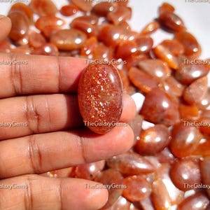 May include: A close-up of a hand holding a large, oval-shaped, sunstone cabochon. The cabochon is a deep orange color with a shimmery, iridescent finish. The background is filled with many smaller, similar sunstone cabochons.