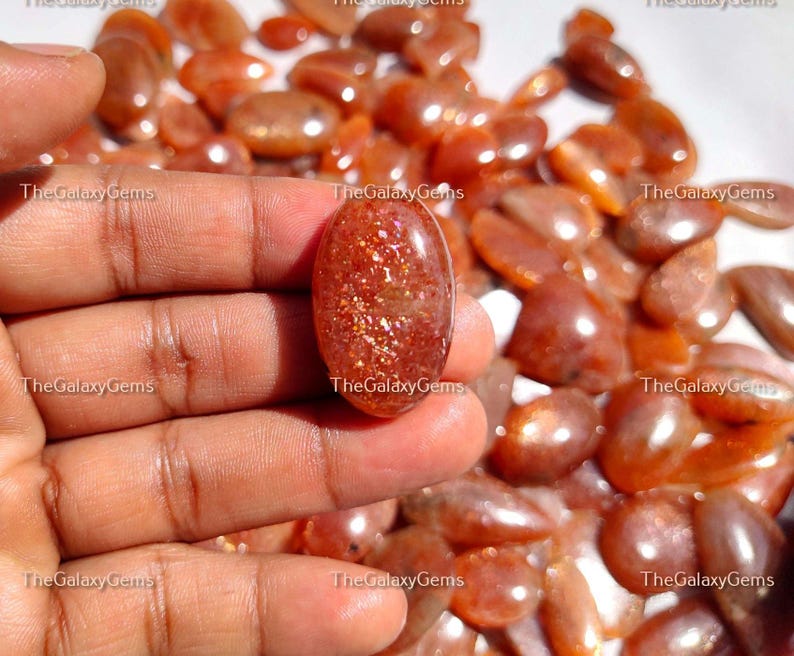 May include: A close-up of a hand holding a large, oval-shaped, sunstone cabochon. The cabochon is a deep orange color with a shimmery, iridescent finish. The background is filled with many smaller, similar sunstone cabochons.