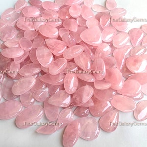 May include: A large pile of pink rose quartz gemstone cabochons in various sizes and shapes. The stones are smooth and polished, with a translucent appearance.