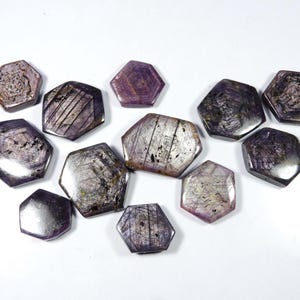May include: A collection of hexagonal gemstones in shades of purple, gray, and silver. The gemstones have a polished surface with visible internal patterns and striations. The arrangement is on a white background, showcasing the variety of shapes and colors.