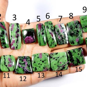 May include: A collection of sixteen polished zoisite gemstones, each displaying a unique pattern of green and magenta hues. The stones vary in shape, including rectangular and square cuts, and are held in a hand.