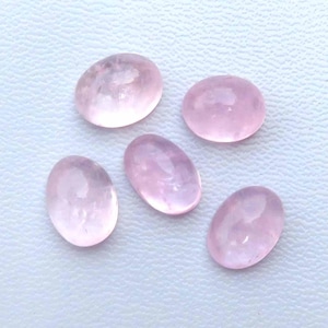 Puede incluir: Cinco piedras preciosas ovaladas de cuarzo rosa. Las piedras son de color rosa suave y tienen una superficie lisa y pulida. Las piedras preciosas están dispersas sobre una superficie blanca.