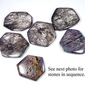 May include: Six polished, irregularly shaped gemstones in shades of gray, purple, and brown are arranged on a white surface. The stones have a reflective, metallic appearance with visible internal patterns. Text at the bottom reads, "See next photo for stones in sequence."