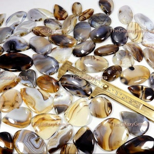 May include: A collection of 25 agate cabochons in various shades of brown, white, and gray. The cabochons are oval shaped and have a smooth, polished finish. The cabochons are arranged in a random pattern on a white surface. TheGalaxyGems is printed on the surface.