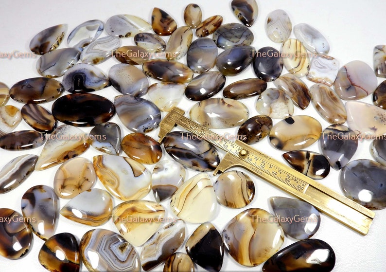 May include: A collection of 25 agate cabochons in various shades of brown, white, and gray. The cabochons are oval shaped and have a smooth, polished finish. The cabochons are arranged in a random pattern on a white surface. TheGalaxyGems is printed on the surface.