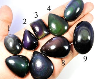 May include: A collection of nine polished obsidian gemstones in various shapes, including oval, teardrop, and rectangular. The stones display iridescent colors of green, purple, and black, with a glossy finish. The gemstones are numbered 1 through 9.