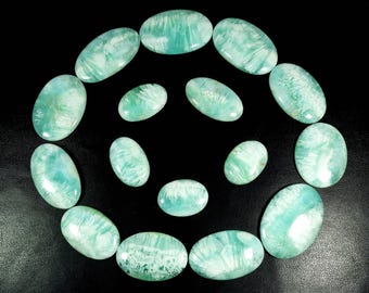 May include: A selection of oval gemstones of varying sizes, arranged in a circular pattern against a black backdrop. The stones exhibit a mix of light blue and white tones, with distinctive internal patterns. The gemstones are polished and smooth.