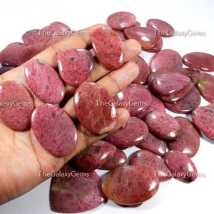 May include: A collection of pink and red oval-shaped gemstone cabochons. The stones have a smooth, polished surface and a speckled pattern.