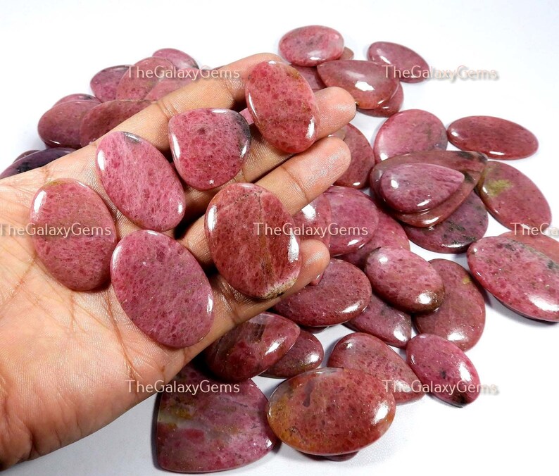 May include: A collection of pink and red oval-shaped gemstone cabochons. The stones have a smooth, polished surface and a speckled pattern.