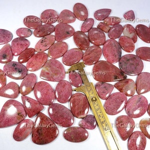 May include: A collection of 30 pink and white gemstone cabochons, each with a unique pattern and shape. The stones are arranged on a white surface with a ruler for scale.