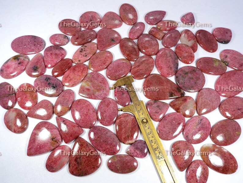 May include: A collection of 30 pink and white gemstone cabochons, each with a unique pattern and shape. The stones are arranged on a white surface with a ruler for scale.