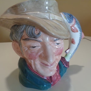 May include: A ceramic pitcher shaped like a man wearing a brown hat and holding a fish. The man has a mustache and a beard, and he is smiling. The fish is blue and white, and it is holding the pitcher's hat.