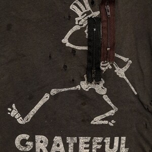 May include: A black t-shirt with a distressed look featuring a white skeleton graphic with a top hat and the text "GRATEFUL DEAD". The shirt has three zippers attached to the skeleton graphic.